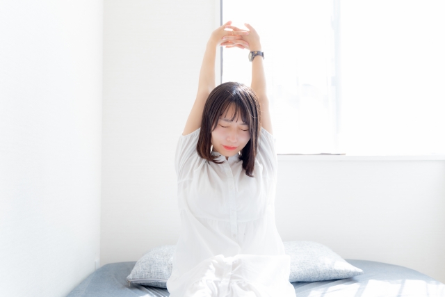 朝の自然光を浴びながら目覚め、ベッドの上で気持ちよく伸びをする若い日本人女性。白いナイトガウンと黒髪が印象的で、睡眠の質やクロノタイプ（朝型・夜型）に関心のある健康志向の女性のイメージに合う。