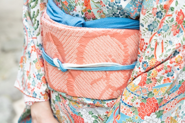 華やかな花柄の着物を身にまとった若い女性の腰元に、繊細な刺繍と柔らかな重なりが美しい淡いピンクの帯が優雅に結ばれている様子。伝統的な和装の中に可憐さと気品が同居し、日本文化における「装い」と「身体表現」の意味を静かに物語る一枚。摂食行動や身体意識を取り巻く文化的背景を探る本コラムの文脈において、「飾ること」「魅せること」の象徴としての装いを視覚的に示している。