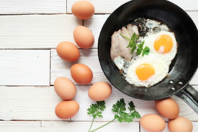 朝食のイメージ：木製テーブルに置かれたフライパンの中に目玉焼きと少量の肉料理、彩りのためのハーブが添えられている。高タンパクでバランスの取れた朝食の一例として、体重管理や食欲調整に役立つ食品を表現。朝ごはんの重要性や栄養構成に関するコラム内容に関連した装飾画像。