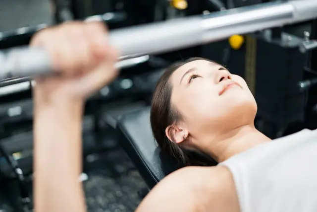 ベンチプレスを行う笑顔の女性アスリート。月経周期やホルモンバランスを考慮したトレーニングと栄養戦略の重要性を象徴するシーン。