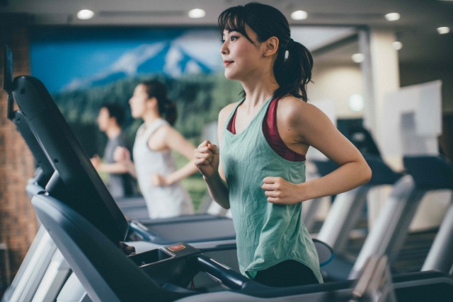 健康的でスリムな体型の日本人女性がジムのトレッドミルでランニングをしている様子。タンクトップから引き締まった二の腕が見え、運動習慣によって維持された筋肉量の重要性を示唆。若い女性のボディイメージ、筋肉の健康、美容とフィットネス、サルコペニア予防といった本記事のテーマに関連する象徴的なイメージ。