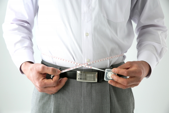 オフィスワーカー風の男性がシャツの上からウエストメジャーで腹囲を測定している様子。わずかに膨らんだお腹が見える。断続的断食（インターミッテント・ファスティング）やカロリー制限が注目される背景には、こうした日常的な内臓脂肪の増加があることを示唆するイメージ。