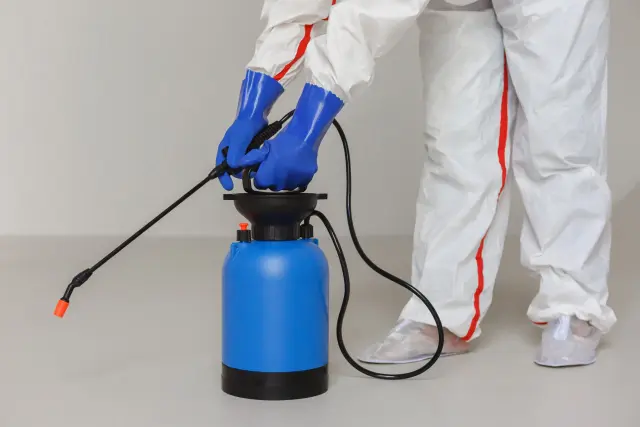 青い防護手袋と作業着を着用した人物が、大型の農薬噴霧タンクを準備している様子。GMO（遺伝子組換え作物）に関連する農薬使用の現場を示すイメージで、農業現場における安全対策や化学物質との関係性を視覚的に表現