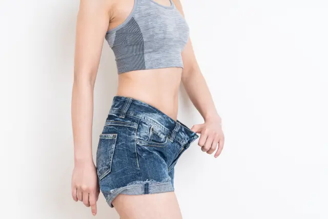 減量に成功した女性が、ゆるくなったホットパンツのウエスト部分を親指で引っ張り、明らかにサイズが合わなくなったことを示しているシーン。精密栄養や運動習慣を通じた脂肪減少と筋肉の引き締めを視覚的に表現。