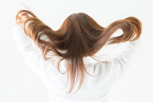 女性が艶やかな髪を肩越しにふわりと翻す瞬間。重みのあるしなやかな髪が滑らかに流れ、健康な毛髪の美しさとボリューム感を印象づける一枚。ヘアケアや毛髪再生の理想像を体現するシーンです。