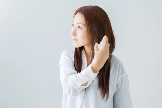 長く艶やかな髪の女性が、髪にミストを吹きかけながら遠くを見つめる。ヘアケア習慣を楽しんでいる様子が伝わる、美髪ケアや保湿ミスト使用の理想的なシーン。