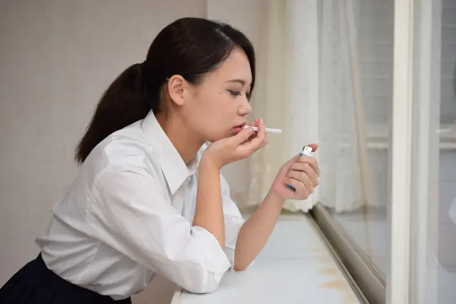 ブラウス姿で髪をひとつ結びにした女性が窓辺に寄りかかり、左手に持つライターを見つめながらタバコを唇に当てている様子。喫煙習慣と健康リスクを想起させるイメージ。