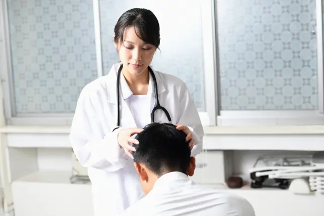 優しい表情を浮かべた女性医師が、細くしなやかな指先で患者の頭皮を丁寧に診察している様子。穏やかな笑顔をたたえながら、慎重かつ思いやりのある態度で頭髪と頭皮の状態を確認しているシーン。