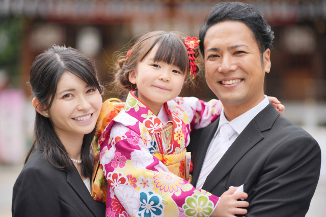 カラフルな花柄着物を着た幼児の娘と、それを抱き笑顔を交わす両親の幸せな家族の姿