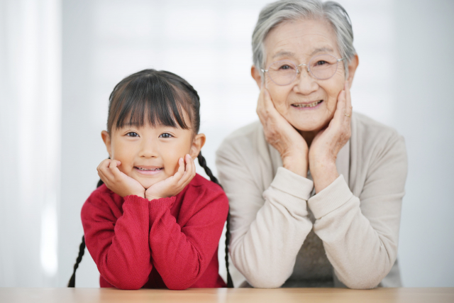 同じポーズで並んで座る祖母と孫娘。木製のテーブルにひじをつき、そっくりな笑顔を浮かべながら寄り添う二人。髪色の違いが印象的で、祖母の白髪と孫娘の黒髪が世代のつながりを象徴する温かな光景。