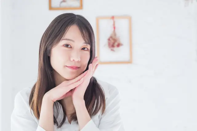 両手を優しく重ね、ほほえみを浮かべる女性が、手元に頬をそっと添える姿。純白の背景と衣装が、胸元まで流れる深いブラウンの髪と瞳の輝きを引き立てる。自分らしさを表現するヘアスタイルと柔らかな表情の調和を象徴するイメージ。