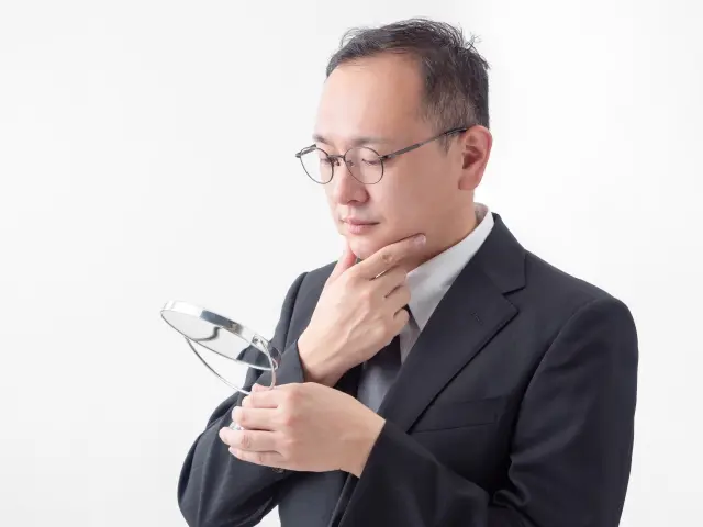 生え際の後退に悩む中年男性が手鏡を見つめながら顎に手を添え、薄毛治療を始めるべきか真剣に考えている様子。AGA治療や植毛の意思決定に悩む男性のイメージ。
