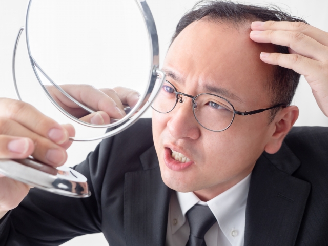 手鏡を震える手で持ちながら、生え際の後退部分に触れ、不安そうな表情を浮かべるビジネスマンの様子