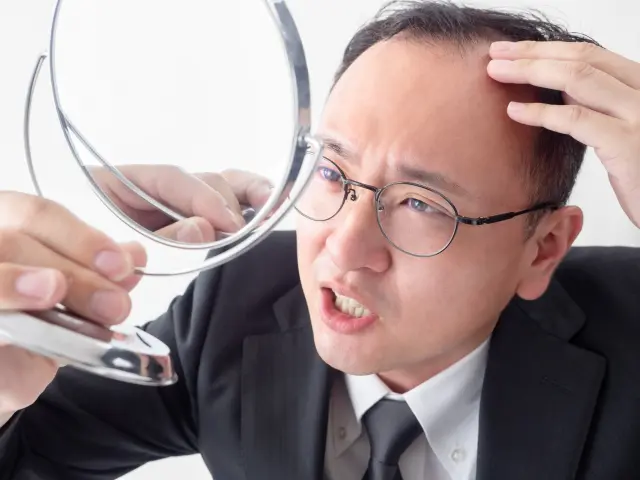 手鏡を震える手で持ちながら、生え際の後退部分に触れ、不安そうな表情を浮かべるビジネスマンの様子