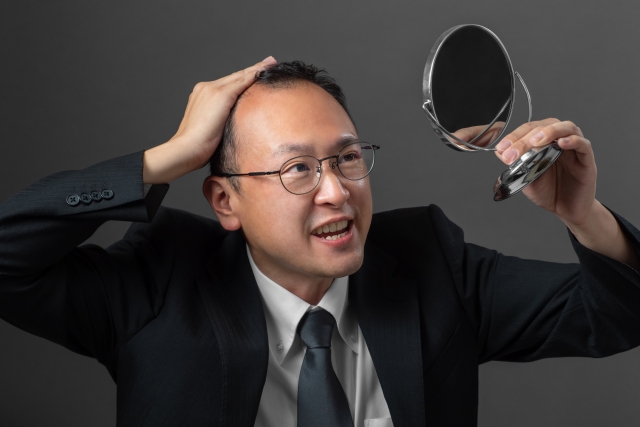 スーツ姿で眼鏡をかけた男性が手鏡を見上げ、髪の薄さに悩むような表情を浮かべている様子