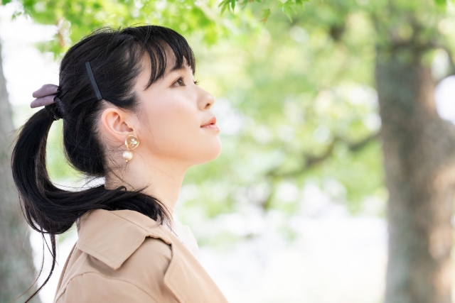 木漏れ日が差し込む中、つややかなポニーテールの女性が上を見上げる姿。柔らかな光が顔立ちを優しく照らし、外見の変化に対する期待や、髪型が与える心理的影響を象徴するような静かな瞬間。