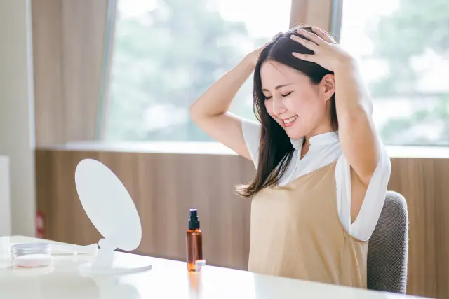 窓辺の鏡の前で、頭にそっと手を添える女性。指先が髪を優しく包み、笑顔に満ちた表情が陽の光に照らされる。心地よい朝のひとときとともに、頭皮と髪の健康を感じる瞬間。