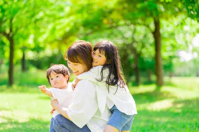 緑豊かな公園で、母親と幼い息子・娘が寄り添って笑顔を見せる親子のひととき。遺伝による髪質の違いや家族の外見的特徴が自然と伝わる場面は、髪の色・太さ・クセが見た目の印象に与える影響を表すイメージ