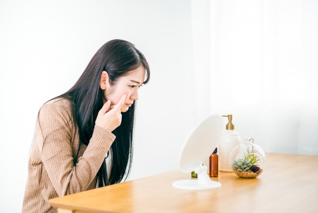 鏡の中の自分を不満げに見つめる女性。指先は頬に添えられ、気になるニキビを見つけた様子。肌トラブルに悩む瞬間こそ、スキンケアや生活習慣の見直しが必要なサイン。