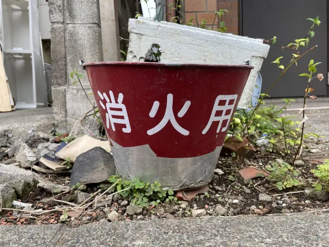 赤い塗装が剥がれ、やや錆びた『消火用』と書かれたバケツが、ゴミや乾いた地面に囲まれて放置されている様子