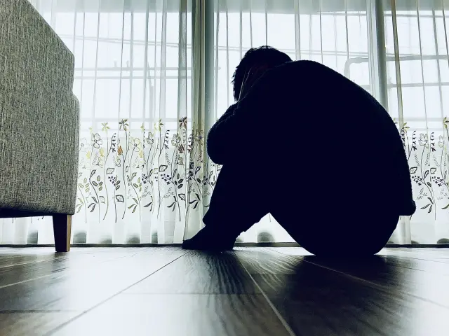 床に座り、膝を抱えてうずくまる若い男性のシルエット。花柄の白いレースカーテンから柔らかな光が差し込むが、彼とその不安な心を照らすことはない。若年層における薄毛・脱毛による自尊心の低下を象徴し、すぐに植毛に頼るのではなく、他の解決策を慎重に検討する重要性を伝えている。