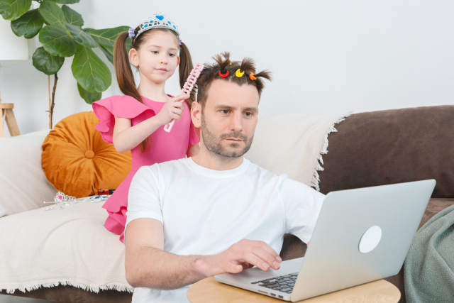 パソコンで仕事をする父親の髪を楽しそうに触る幼い娘の様子。家族のふれあいや父親の髪への関心が伝わる温かい日常の一コマ。薄毛や年齢変化をめぐる家族の視点を象徴。