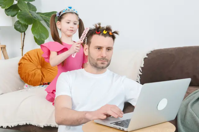パソコンで仕事をする父親の髪を楽しそうに触る幼い娘の様子。家族のふれあいや父親の髪への関心が伝わる温かい日常の一コマ。薄毛や年齢変化をめぐる家族の視点を象徴。