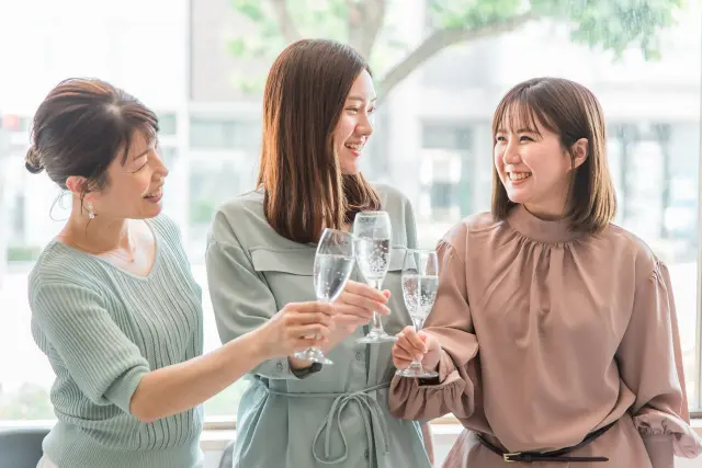 透明なドリンクで乾杯する女性3人のイメージ写真。明るい雰囲気の中、立ったままグラスを掲げて笑顔を交わすシーン。外見の変化やライフスタイルの前向きな変化を象徴するイメージとして使用可能。