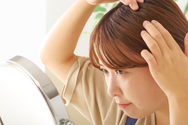 鏡の前で中年女性が指先で頭頂部をなぞりながら髪の状態を確認している。表情には満足感がなく、薄毛や抜け毛に対する不安を感じさせる。頭皮チェックやヘアトラブルの悩みを象徴するシーン。