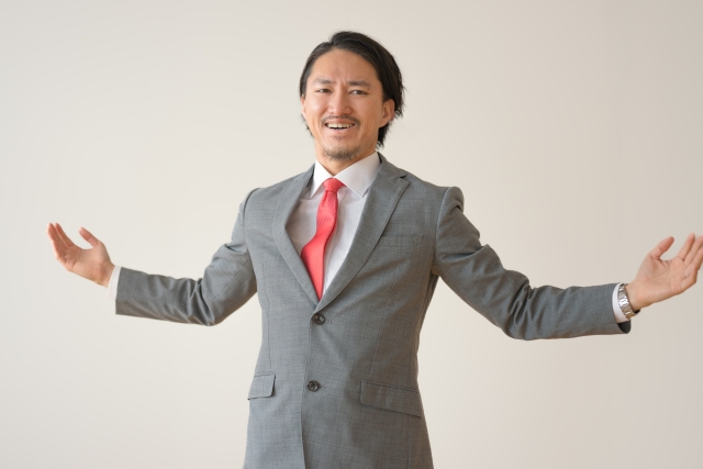 スーツ姿のビジネスマンが両腕を大きく広げ、笑顔でポジティブな印象を与えているイメージ写真。成功、歓迎、自信、開放感を象徴するビジネスシーン向けのビジュアル素材。