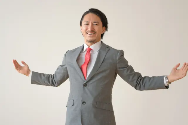 スーツ姿のビジネスマンが両腕を大きく広げ、笑顔でポジティブな印象を与えているイメージ写真。成功、歓迎、自信、開放感を象徴するビジネスシーン向けのビジュアル素材。