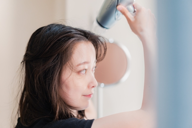 鏡を見つめながら髪をドライヤーで乾かす女性。髪は濡れて艶のある黒髪で、うるおいとハリが感じられる。健康的な美髪ケアや濡れ髪の正しいドライ方法を示す理想的なシーン。