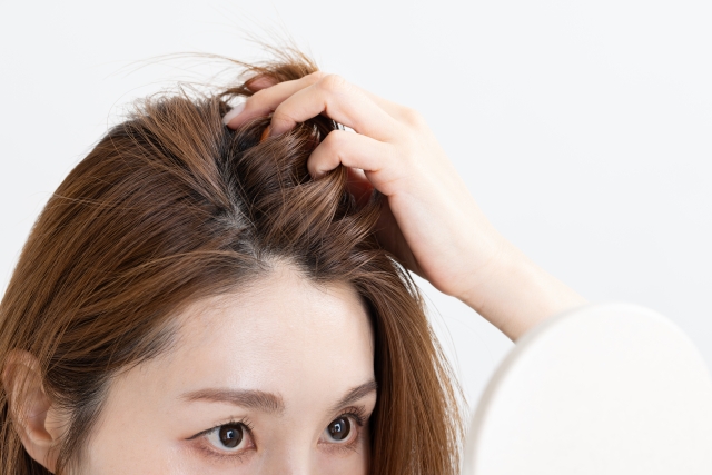 鏡越しに頭皮を見つめる女性。髪をかき上げ、薄くなった分け目や地肌を真剣に確認している。薄毛の悩みは深刻だが、「きっと何か方法がある」と希望を捨てずに向き合う姿。薄毛対策や毛髪再生に関心のある女性に共感を呼ぶ一枚。