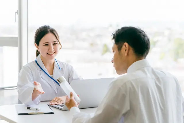 女性医師が穏やかに手を差し出しながら説明し、向かいのデスクで男性患者が薬剤ボトルを手に取り真剣に確認している様子。薄毛治療薬の説明を受けるAGAカウンセリングのイメージ。