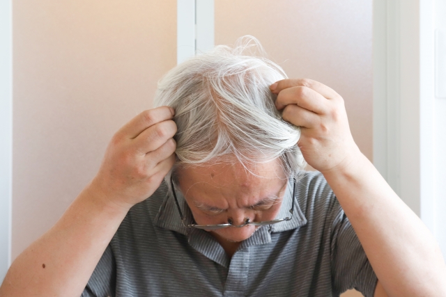 高齢の男性が、頭皮の露出部分を隠そうと白髪を手で整え、髪をかき寄せている様子。薄毛が進行し、頭皮の見える面積が広がっている状況を表現したシーン。