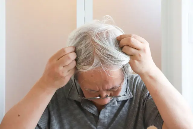 高齢の男性が、頭皮の露出部分を隠そうと白髪を手で整え、髪をかき寄せている様子。薄毛が進行し、頭皮の見える面積が広がっている状況を表現したシーン。