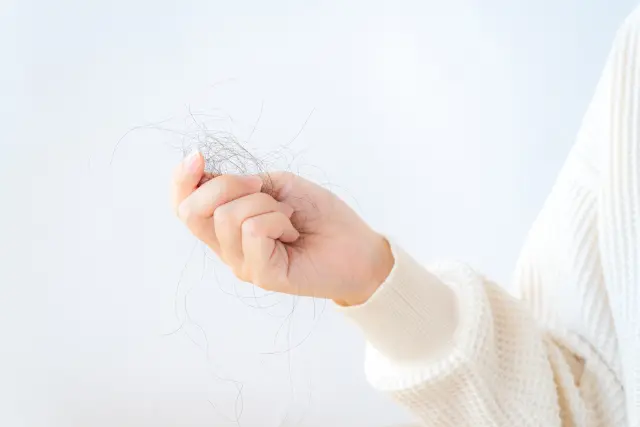 柔らかなニットセーターの袖口から伸びた女性の手が、抜け落ちた細い髪の束を軽く握っている様子。抜け毛や薄毛が進行する不安や恐怖を感じさせる場面。