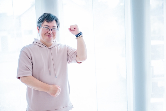 前向きな笑顔を見せながら、左手を軽く握って励ますようなポーズをする中年男性のイメージ写真。自信やポジティブな気持ち、希望を表現したビジュアル素材。