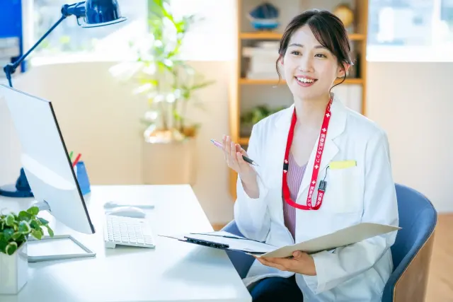 デスクに座りながら広い笑顔を浮かべる女性医師。片手でペンを持ち、もう片方の手にクリップボードを持っている姿が映し出されています。明るく活気ある雰囲気の中で、医療相談とサポートを通じて、髪の悩みや自己像の回復がうまく進むことへの希望を感じさせます。