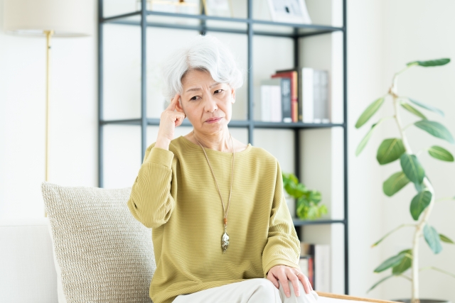 白髪が目立つ高齢女性が、指をこめかみに当てながら床を見つめ、加齢による薄毛と植毛の可能性について思い悩む様子。白髪でも植毛できるのかを真剣に考える姿。