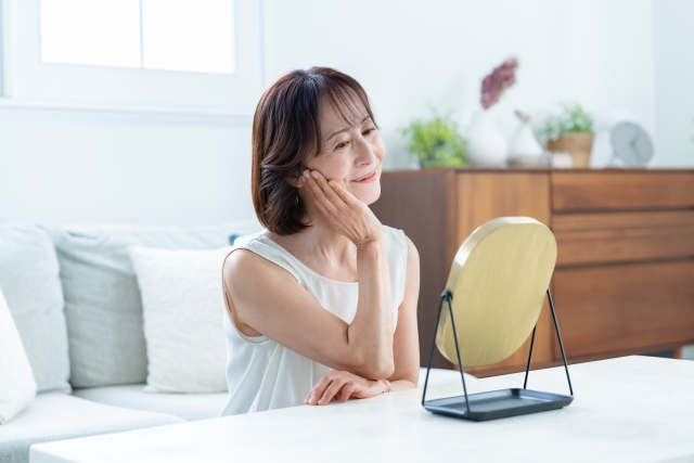 加齢による髪や顔の変化を見つめながら、鏡の前で静かに自分の容姿を見つめる高齢女性。自然な老いを受け入れ、白髪や肌の変化を優雅に受け止めたいと願う様子。