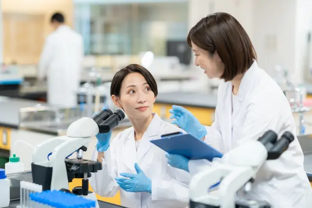白衣を着た2人の女性研究者が実験室で業務中。1人はベンチで顕微鏡を操作し、もう1人はクリップボードを持って議論を交わす医療・研究現場のイメージ|臨床試験や医薬品研究、発毛治療薬の科学的検証に関連する画像素材