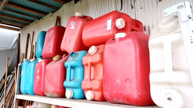 木製の棚に並べられた赤や青の空のガソリン容器。日焼けで色褪せたものも含まれ、プラスチック製の色の違いでは、引火事故を防ぐことはできないというリスクを象徴的に表現した構図。