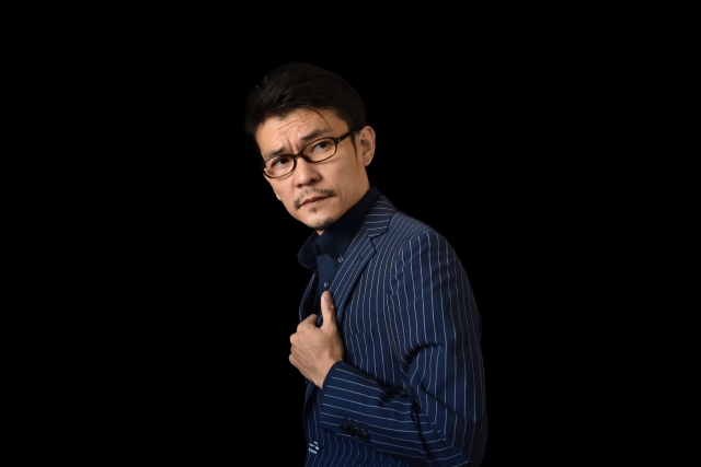 黒い背景の中、濃紺のシャツを着て横を見つめる眼鏡の日本人男性。左腕を胸に添えたポーズと、整った口髭と顎髭が洗練された男らしさを演出