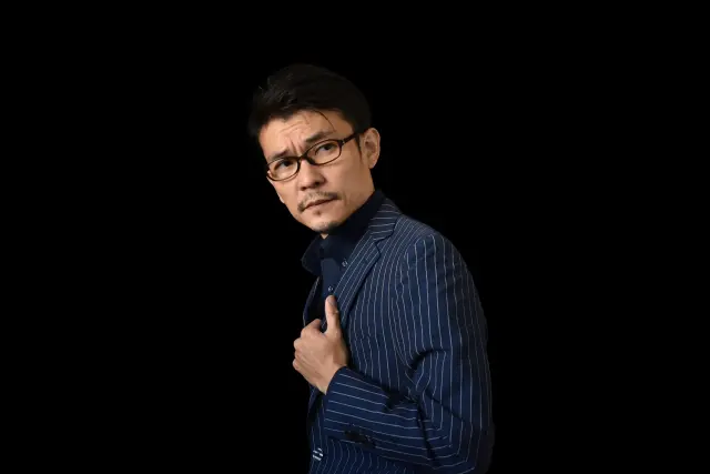 黒い背景の中、濃紺のシャツを着て横を見つめる眼鏡の日本人男性。左腕を胸に添えたポーズと、整った口髭と顎髭が洗練された男らしさを演出