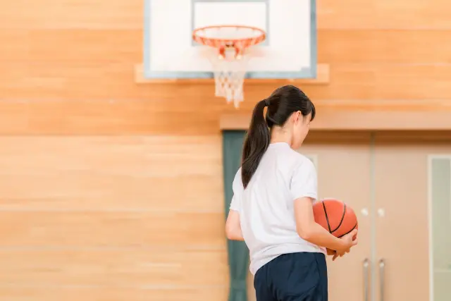 バスケットボールコートに立つ若い女性。白いTシャツとブルマー姿で、右腕にオレンジ色のバスケットボールを抱えながら背中を向けて立つ。スレンダーな背中にかかる艶やかなポニーテールが、若さと健康的な活力、そして美しい髪の象徴として印象的なイメージ