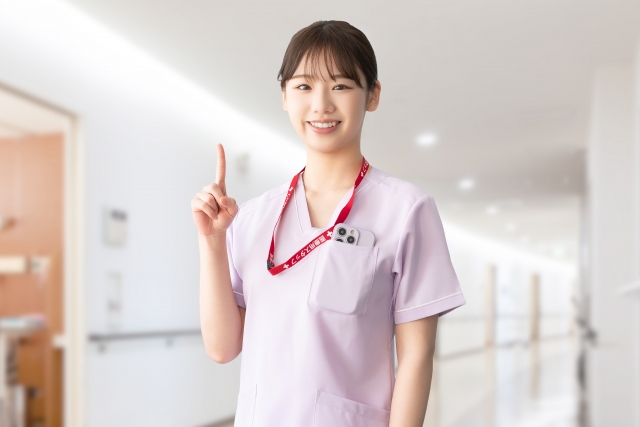 淡いピンクの制服を着た看護師が、人差し指を立てた可愛らしいポーズで微笑む様子。安心感と親しみやすさがにじみ出る笑顔が、患者への丁寧な対応や信頼できる医療サービスを感じさせるワンシーンです。