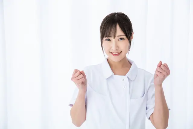 白衣をまとい、両手を軽く握ったポーズで温かく迎える女性医療スタッフ。笑顔と励ましのボディランゲージで、患者との信頼関係構築の重要性を表現