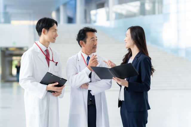 白衣を着た2人の男性研究者が、黒いスーツ姿の女性と和やかに会話する場面。手にはメモやクリップボードを持ち、薄毛治療や遺伝子研究に関する専門的な意見交換が前向きに進んでいる様子を描写したイメージ。