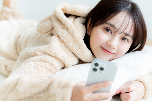 ふわふわのウールジャケットを着た若い女性がマットレスの上に横たわり、スマートフォンを片手に持ちながら可愛らしく微笑んでいます。髪の悩みや自己像に対する不安を抱えている方々が、自分らしさを取り戻すために前向きな気持ちを持つことの大切さを象徴しています。